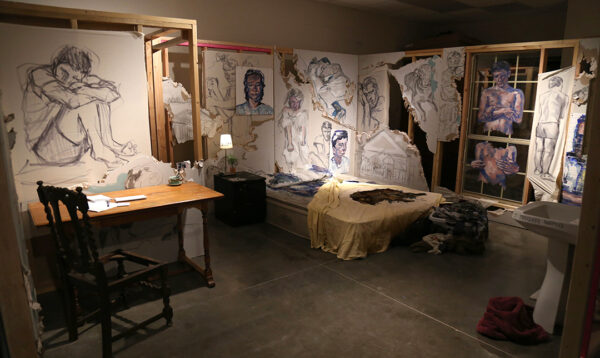 A photograph of an installation by Elijah Ruhala featuring portraits set into a built scene resembling a small one room apartment.