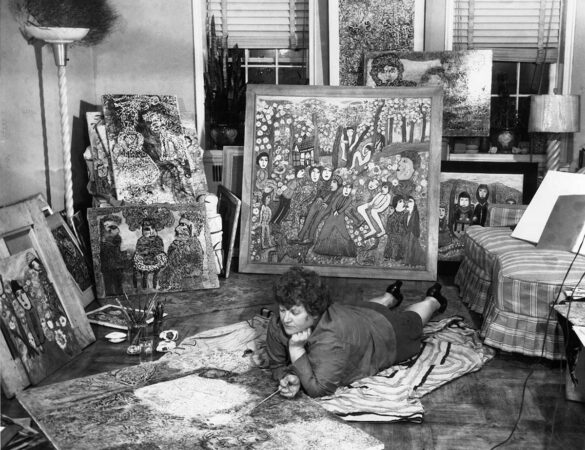 A woman lays on the floor of her apartment painting a canvas. She is surrounded by other paintings and art supplies.