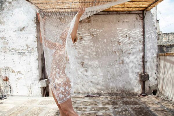 A nude woman stands behind a lace textile hung outside on the roof an Italian house on a bright day.