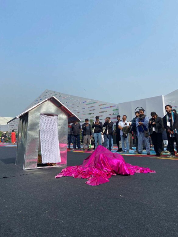 A bright pink cloth covers an artist during a performance next to a small sculpture of a house.