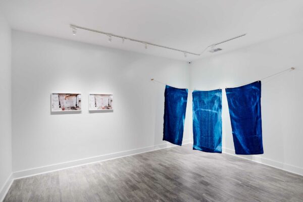 Two photographs hang on the wall of a gallery, while three cyanotype textiles hang from the ceiling.