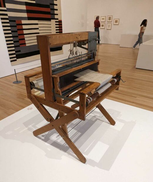 A weaving loom once owned by Anni Albers sits on a plinth in the foreground while on the wall in the background hangs one of Alber's weavings.
