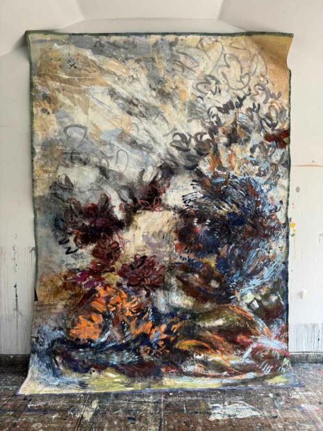 A large scale, unstretched, abstract painting hangs on the wall with the bottom portion laying on the ground.