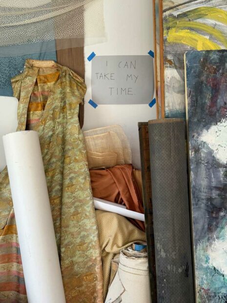 Rolls of paper, fabric, and other art materials lean against the wall of a studio. Taped to the wall is a message reading "I can take my time."