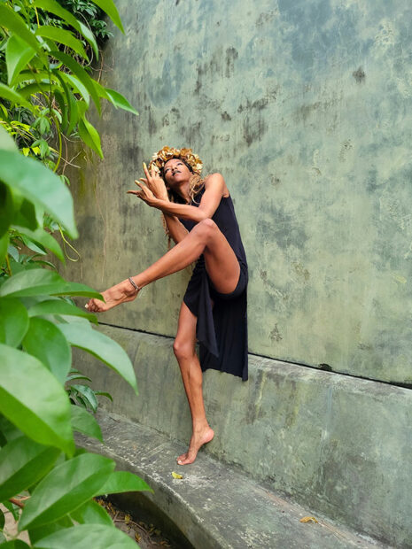 A dancer in a black dress performs outdoors against a concrete wall.