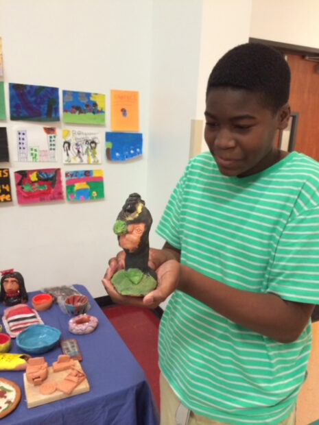 A young man in a green shirt holds a small clay sculpture towards the camera.