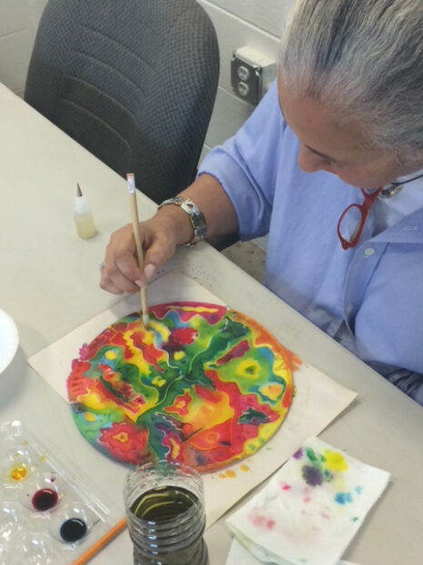 An elderly woman works on a small round painting on a desktop.