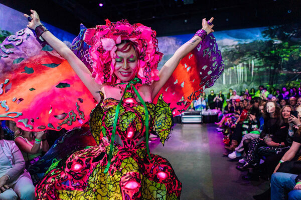 A woman poses for the camera with her arms in the air wearing a brightly colored outfit made from discarded objects.