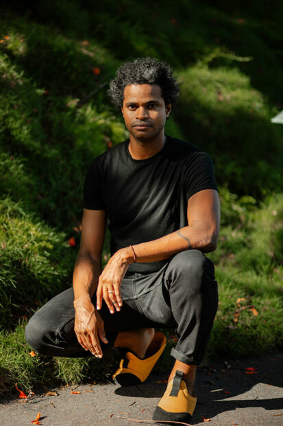 A man in a black t-shirt and jeans takes a knee outdoors and faces the camera for a portrait.