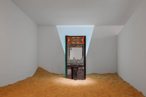 An installation by Violette Bule featuring a claw game machine installed in a room with a dirt covered floor.