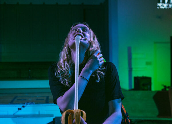 A blonde woman in a black t-shirt performs on stage with a banana prop.