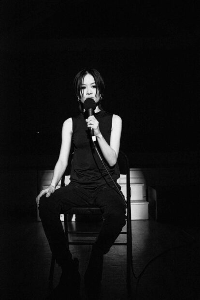 A woman in black clothes sits onstage speaking into a microphone.