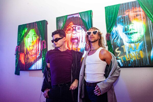 Photo of two men posing in front of three mixed media portraits on a wall