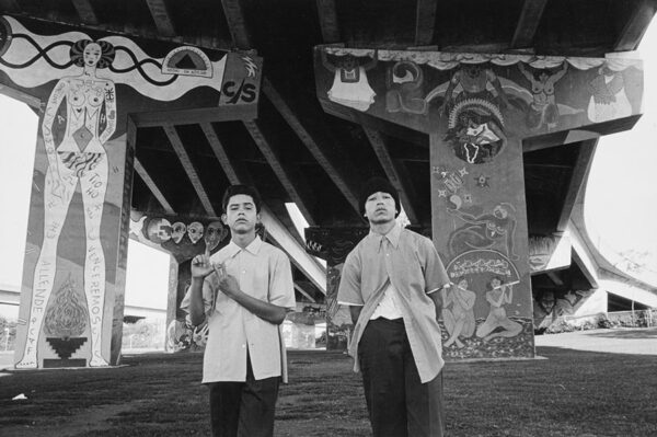 Two young men stand beneath a an overpass decorated with Chicano murals. One throws a gang sign for Vatos Locos. 