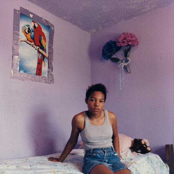 A young woman sits on a bed in a sparsely decorated bedroom, staring into the camera.