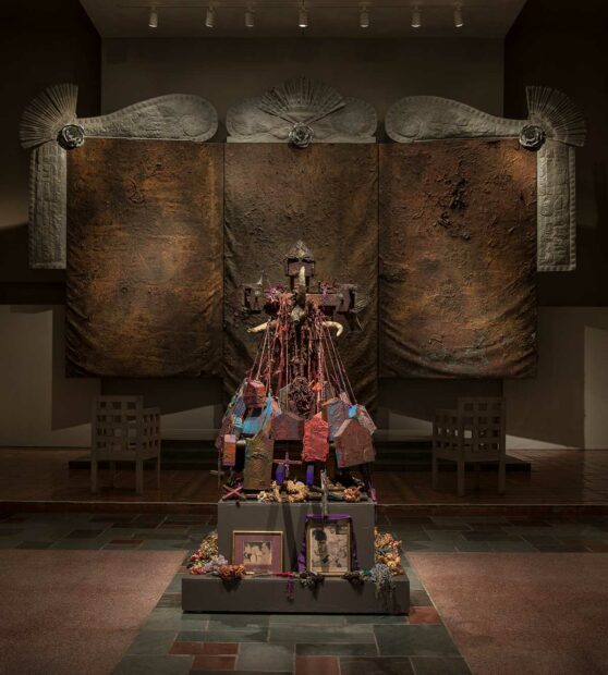 A shrine-like sculpture comprised of framed images and small structures sits before a large wall-sized painting.