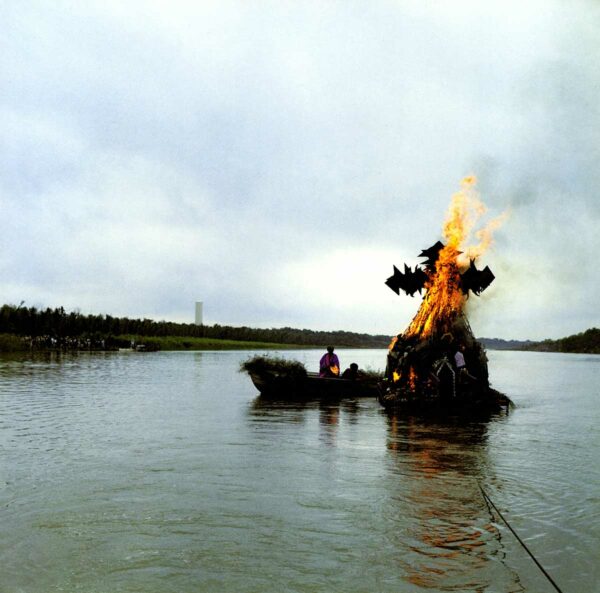 A photograph documenting a performance in a river with a large sculpture on fire and a figure in a boat next to it.