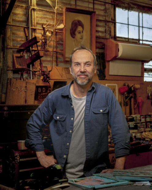 A man stands in a woodshop in a blue button down shirt.