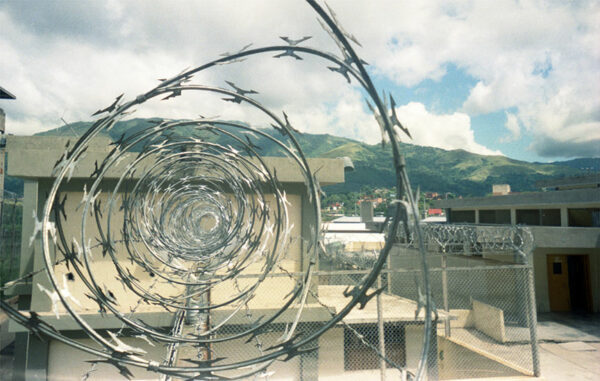 A spool of razor wire sits in the center of a photograph of a prison.