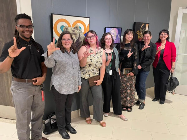 A photograph of a small group of people standing in front of works of art and holding up an "L" symbol with their hands.