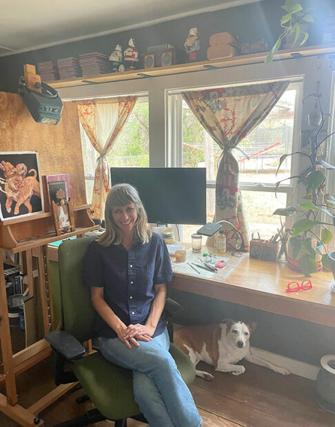 A woman sits at her desk facing the camera with a white dog on the floor and painting and drawing material surrounding her.