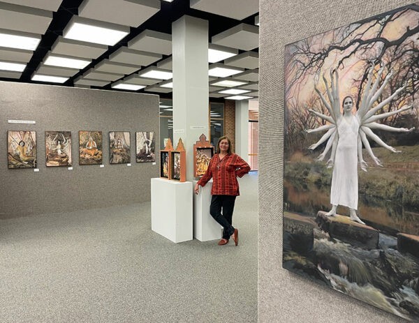 A woman stands in a large gallery with figurative paintings hanging on the wall, looking directly into the camera.