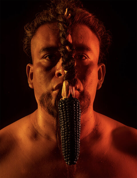 A photograph by Manuel Chavajay featuring a man with braided hair in front of his face and an ear of corn hanging from the end of his hair.