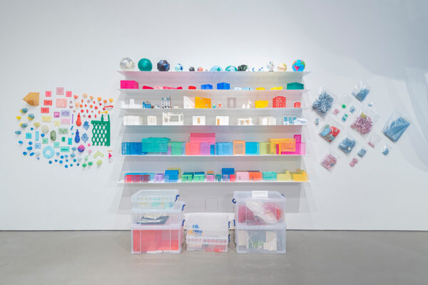 Brightly colored items from the artist's studio are displayed on shelves, the gallery walls, and in clear plastic boxes on the floor of a gallery.