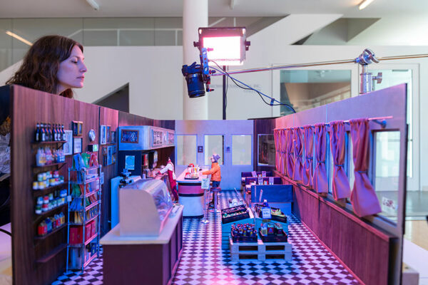 A woman peers over the edge of a miniature model of a diner with a solitary figurine sitting at the counter.