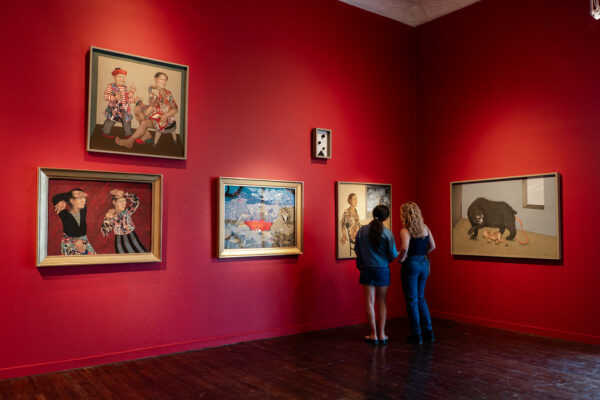 A gallery with red walls displays several surrealist paintings hung salon style.