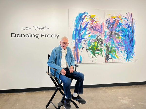 A man sits in fron of a gallery wall displaying a diptych of his non-figurative abstraction.