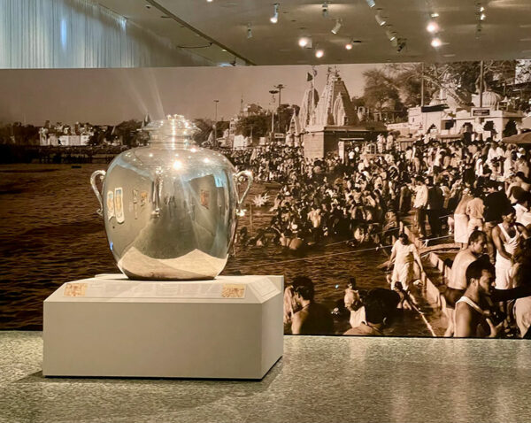 A large silver pot is on display in front of a wall sized image of people in India.