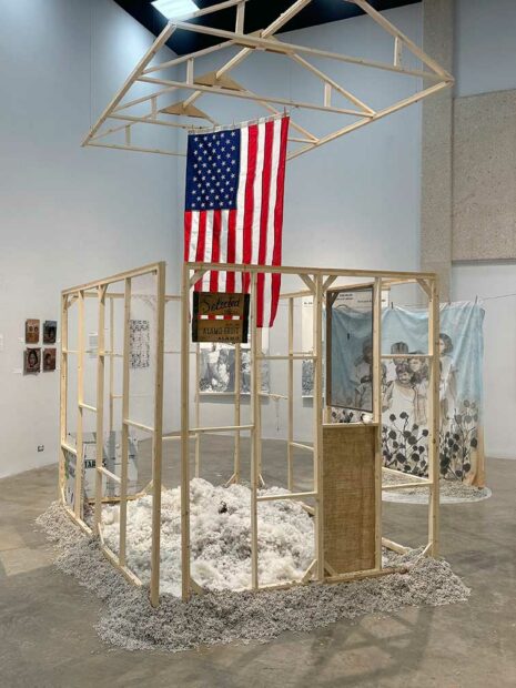 A large installation consisting of a wooden house structure with an American flag, and piles of cotton seed, and cotton