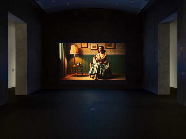 A still image of a woman sitting in an empty room waiting on a phone call, from a video work by Laurie Simmons.