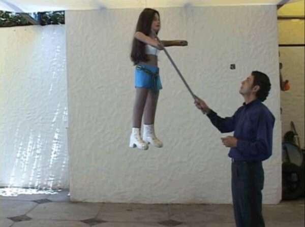 A man stands beneath a piñata in the shape of a woman, touch a stick to her face.