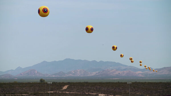 A row of yellow weather balloons with a "watchful eye" design on them float above the U.S. / Mexico border.