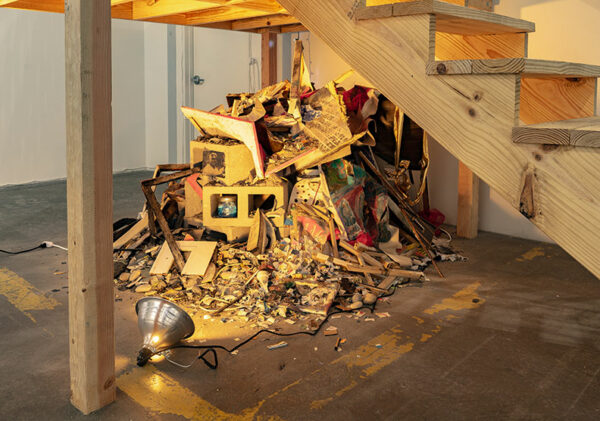 Debris piled up underneath a platform in an art gallery features cinderblocks, photographs, damaged paintings.