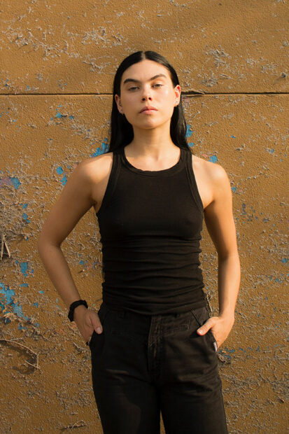 A woman with black hair stands before a brown wall staring into the camera, wearing all black.