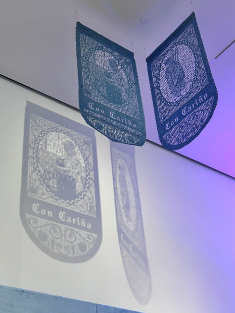 Two blue papel picados hang from the ceiling of a gallery casting a shadow on the walls.