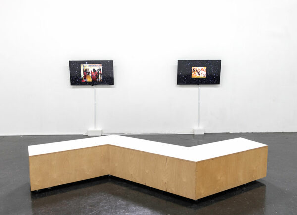 Two monitors installed on a gallery wall before a large bench.