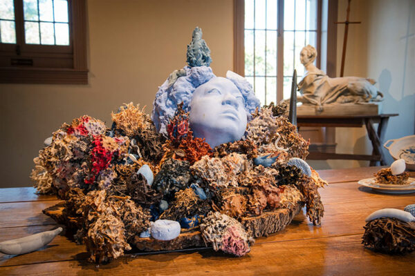 A blue female head stares upward from a pile of dried flowers on a table in a gallery.