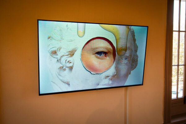 A video plays on a monitor hung on a gallery wall of an image of a classical statue with a model's eye peering through a hole cut in the image.