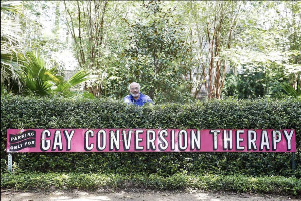 A photograph of artist Robert Rosenberg standing behind a side that reads, "Parking only for Gay Conversion Therapy."