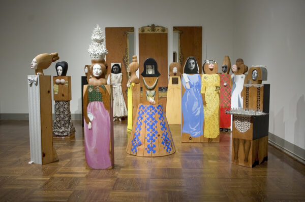 An installation of a work by Marisol featuring 15 freestanding life-size figures and 3 panels.