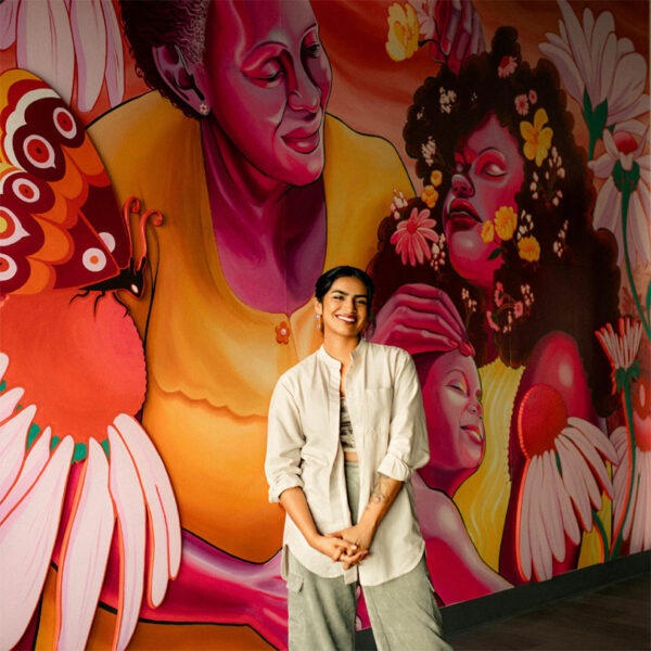 A woman stands in front of a large figurative mural.