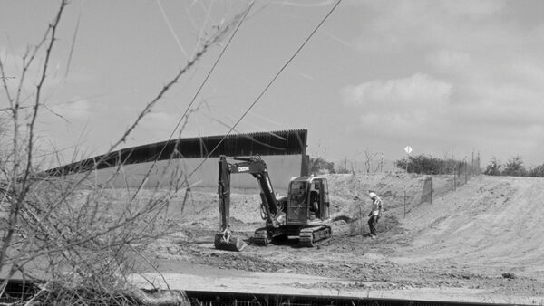 Construction equipment and a lone worker stand before a border wall.