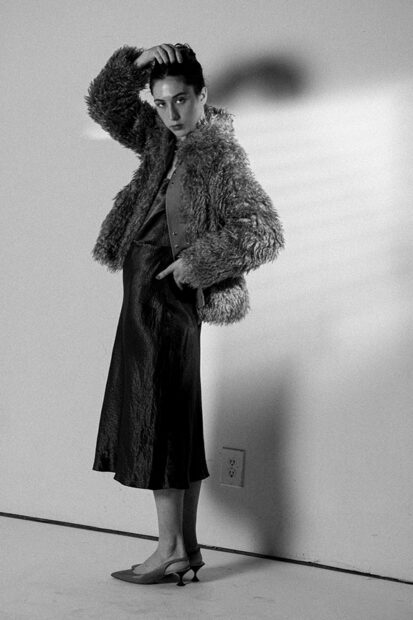 A woman in a fur coat and dress stands with one hand on her head looking directly into the camera.