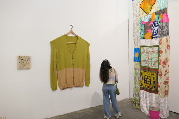 An oversized sweater hangs on an oversized wooden hanger on a gallery wall.