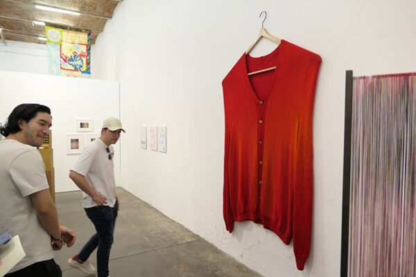 An oversized sweater hangs on an oversized wooden hanger on a gallery wall.