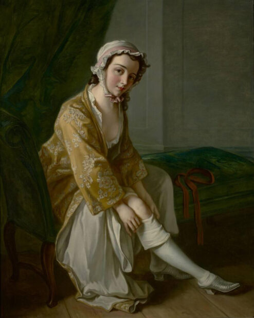 A woman in a dark room pulls up her stocking while looking directly at the viewer.